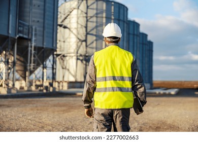Back view of an industry worker walking towards silos with tablet in his hands.