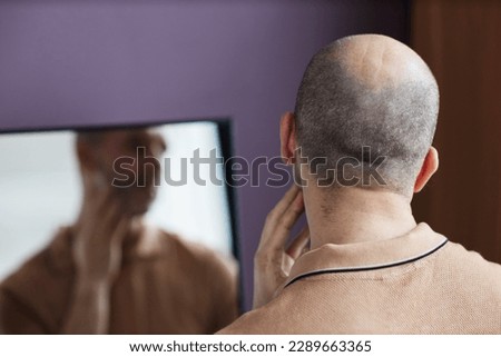 Back view of insecure bald man looking in mirror at home and inspecting face, copy space