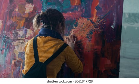 Back View Of An Isolated Caucasian Female Using Her Fingers To Paint On A Big Modern Abstract Canvas, Wearing Dark Overalls And An Orange Sweater, Contemporary Artist Using Her Hands To Paint. 