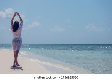 Back View Of Joyful Cute Attractive Young Happy Woman In White Dress With Arms Up On Sand Beach Enjoying Ocean, Summer Breeze And Sound Of The Waves During Vacation. Happy Holiday Travel Concept.