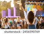 Back view of joyful woman with raised hands enjoys summer music festival. Crowd dances at beach concert, sunset light. Happy fan cheers at outdoor live event. Excited attendee celebrates, vibes.