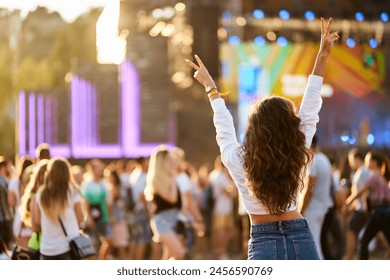 Back view of joyful woman with raised hands enjoys summer music festival. Crowd dances at beach concert, sunset light. Happy fan cheers at outdoor live event. Excited attendee celebrates, vibes.