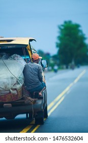 Back View Of Khmer Porter Hangs On Delivery Minivan Transports A Full Load Of Food And Beverage On The Road Of Rural South Cambodia. Motion Blur.