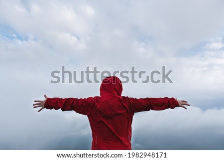 Back view of a kid with extended arms in front of a dramatic sky and wearing a colorful red raincoat. Feeling free and flying sensation concept.