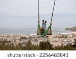 Back view of a kid on the swing. Child enjoying swinging and looking at ocean scene with city along the coast. Traveling to Nazare, Portugal