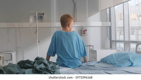 Back View Of Kid Patient Sitting On Hospital Bed Looking Out Of Window. Preteen Sick Boy Sitting On Bed In Hospital Ward. Medicine, Health And Healthcare Service