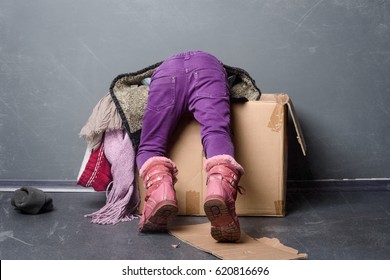 Back View Of A Kid In Shabby Clothes Looking Inside Of A Paper Box, Close-up. Old Boots, A Hat On The Floor, Grey Background. Homeless.