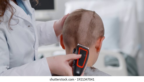 Back View Of Kid Sitting In Hospital Ward And Having Hair Cut Preparing For Brain Surgery. Nurse Or Doctor Shaving Teen Boy Before Chemotherapy Or Operation