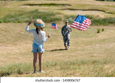 Back View Of Kid In Straw Hat Waving Hand While Holding American Flag Near Father In Military Uniform 