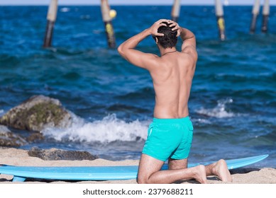 Back view of kneeling man on the beach with his surfboard resting and looking at the sea before surfing with hands on head - Powered by Shutterstock