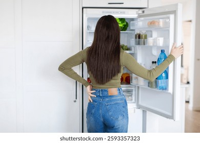 Вид сзади леди, стоящей перед открытым холодильником, полным продуктов питания и выбора ингредиентов для приготовления пищи, свободное место