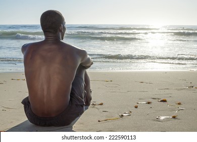 Back view of lean young shirtless African American man sitting on sand while watching waves roll in on beautiful beach under setting sun