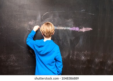 Back view of a little boy painting something on a chalkboard.