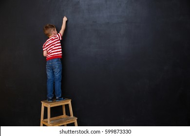 Back view of a little boy standing on ladder and writing with chalk on the backboard in school class