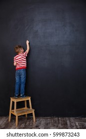 Back view of a little child standing on a chair and drawing on a chalk blackboard
