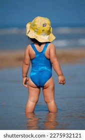 Back view of a little girl in bathing suit and sun hat standing with her feet in the sea under a blue sky 