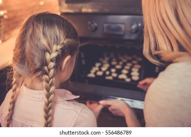 Back View Of Little Girl And Her Mom Putting Cookies Into The Oven While Baking In Kitchen At Home