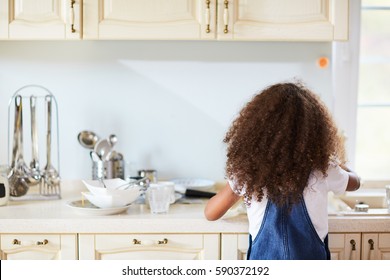 Back View Of Little Girl With Long Curly Hair Standing At Kitchen Sink And Washing Dishes After Family Lunch