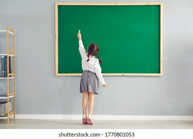 Back view of little girl writing on blackboard with chalk at classroom. Smart first grader answering on lesson at school - Powered by Shutterstock