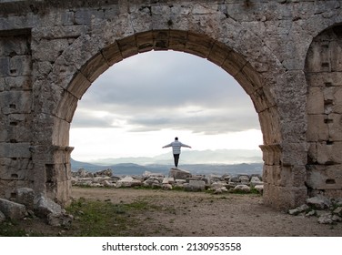 Back View Of Lonely Man Open Arms On Rocks. He Standing In Front Of Old, Stone Entrance. Cloudy Sky, Sunlights And Mountains View. Amazing Landscape Scene. Travel By Alone. Achievement Concept.