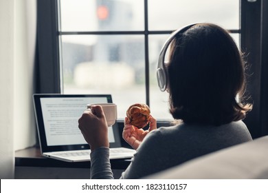 Back View Of Lonely Woman Enjoying Having Breakfast With Cup Of Coffee And Croissant, Listenning Music In Headphones Working On Laptop Sitting Near Window In Cafe. 