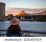 back view of a long hair woman in a hat watching the sunset over brisbane city; city reach boardwalk with amazing view of large skyscrapers by brisbane river, australia	

