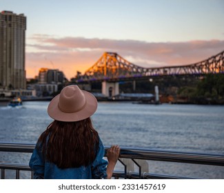back view of a long hair woman in a hat watching the sunset over brisbane city; city reach boardwalk with amazing view of large skyscrapers by brisbane river, australia	
					
