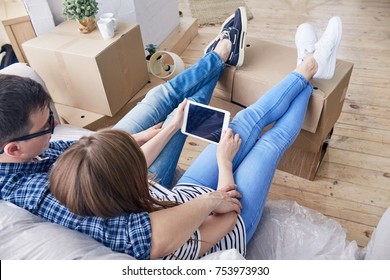 Back View Of Lovely Young Couple Watching Their Favorite TV Series On Digital Tablet And Sitting On Cozy Sofa While Taking Break From Packing Stuff In Moving Boxes.