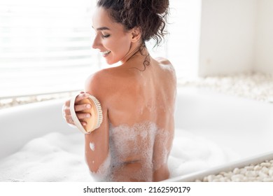 Back View Of Lovely Young Woman Making Dry Brush Lymphatic Massage, Scrubbing Her Skin In Bubbly Bath At Home. Pretty Millennial Lady Taking Care Of Her Body In Bathtub At Hotel