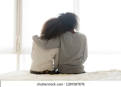 Back View Of Loving African American Mother Hug Teen Daughter Sitting On Bed, Caring Black Mom Embrace Child, Relaxing In Bedroom Looking In Window, Parent Comfort Teenager Caressing At Home