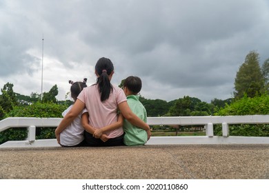 Back View Of Loving Asian Mother Hug Her Kids Sitting On Bridge, Caring Black Mom Embrace Child, Relaxing Looking To Black Cloud, Parent Comfort Child Caressing.