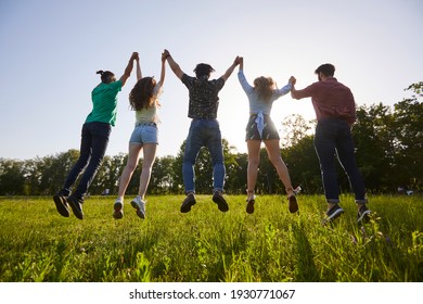 Back view low angle of unrecognizable group of friends holding hands in moment of jumping above ground on green meadow while enjoying summer holiday