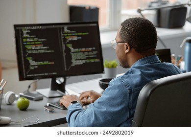 Back view of male African American senior IT specialist in glasses code programming on computer while sitting at desk with green fresh apple at hand in office, copy space - Powered by Shutterstock