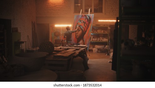 Back View Of Male Artist Painting Portrait Of Black Woman On Easel Against Lamp During Work In Spacious Workshop