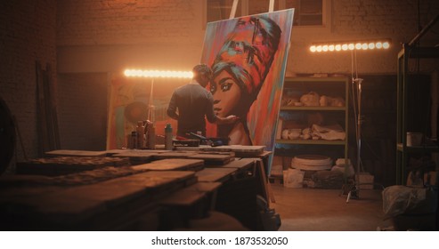 Back View Of Male Artist Painting Portrait Of Black Woman On Easel Against Lamp During Work In Spacious Workshop