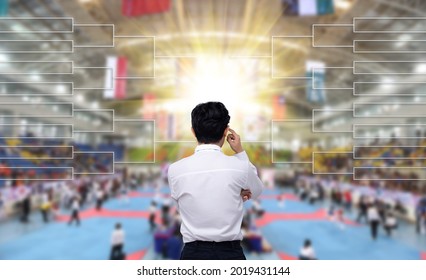 Back View Of Male Coach In White Shirt Watching Schedule Tree Chart Or Timetable So He Can Plan Strategy Of Taekwondo, Karate, Judo Fighting On Quarter Final Match In International Stadium