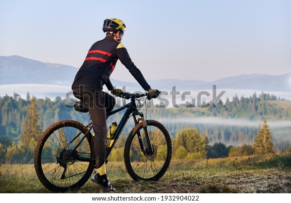 Back view of male cyclist in cycling suit\
standing with bike with coniferous trees and hills on background.\
Man bicyclist enjoying bicycle ride in mountains. Concept of sport,\
biking, active leisure.