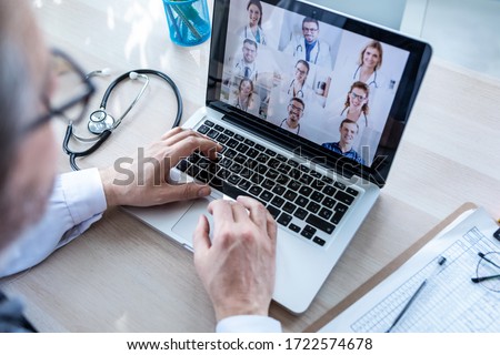 Back view of male doctor having video conference on laptop with his colleagues at home.