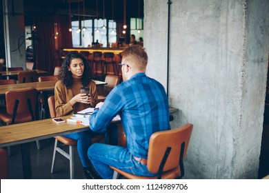 Back view of male employee making interview for dark skinned female student discussing cooperation details, professional colleagues communicating with each other in cafeteria during coffee break