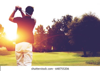 Back View Of Male Golfer Following Through From A Swing With Beautiful Sunset Light On Fairway. Mature Man Practicing His Swing On The Golf Course. 