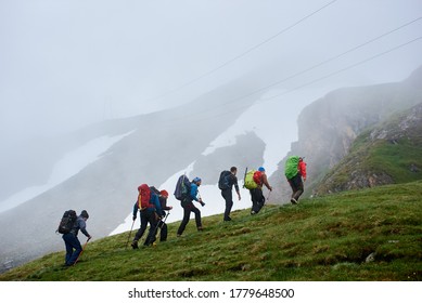 Back view of male hikers with backpacks climbing grassy hill. Young tourists walking uphill in mountains with foggy cliff on background. Concept of hiking, travelling and backpacking. - Powered by Shutterstock