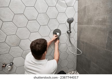Back view of male plumber installing wall mounted shower system in apartment under renovation. Man repairing metal shower faucet handle while working on bathroom renovation. Plumbing works concept.