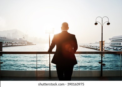 Back view of a male proud CEO is resting after work day, while is standing outdoors against sea port in evening. Silhouette of a young successful businessman in black suit is enjoying landscape