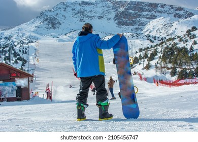 Back view of male snowboarder wearing blue jacket, navy blue pants standing with snowboard in one hand and enjoying alpine mountain landscape - snowboarding concept.
