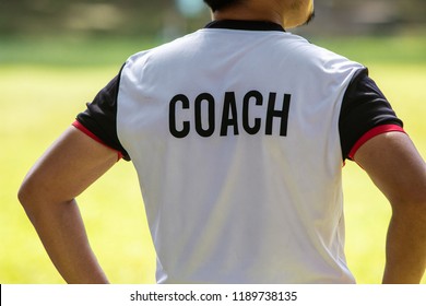 Back View Of Male Soccer Or Football Coach In White Shirt With Word COACH Written On Back, Standing On The Sideline Watching His Team Play, Good For Sport Or Coaching Concept