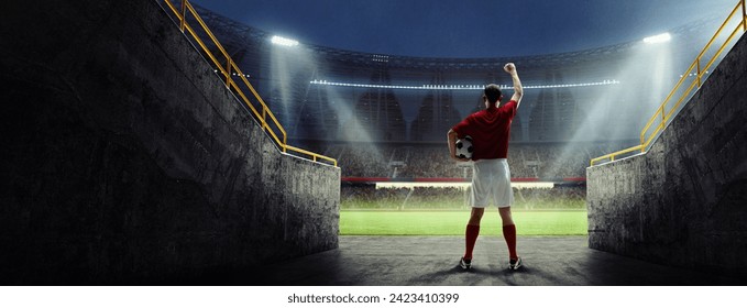 Back view of male soccer player in red jersey standing with ball, raising fist up, facing stadium crowd. Night game atmosphere. Motivation to win. Live sport event, championship and tournament concept - Powered by Shutterstock