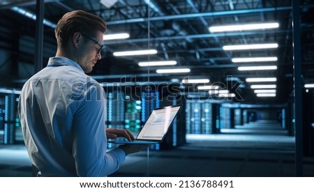 Back View of Male Specialist Using Laptop in Big Data Center Office. High Speed Data Transfer, Server Transfer, Technology Science Breakethrough, Progress, Innovation Concept