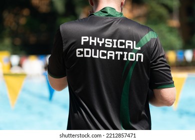 Back view of male swimming coach, wearing PHYSICAL EDUCATION shirt, working at an outdoor swimming pool - Shutterstock ID 2212857471