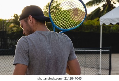 Back View Of Male Tennis Player Outdoors, A Man Wearing Cap Holds Tennis Racquet On His Shoulder.