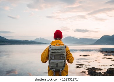 Back view of male tourist with rucksack standing on coast in front of great mountain massif while journey.  Man traveler wearing yellow jacket with backpack explore Scandinavia nature. Wanderlust